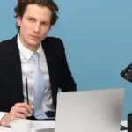 A focused businessman in a suit works at his tidy desk with a laptop, plant, and lamp.