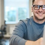 Happy man wearing glasses working remotely on laptop in modern office environment.